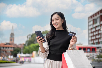 Asian woman or office worker standing outdoor looking at phone holding credit card and shopping bags