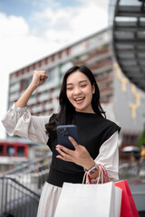 Asian woman office worker standing carrying shopping bags and raising a fist while looking at phone.