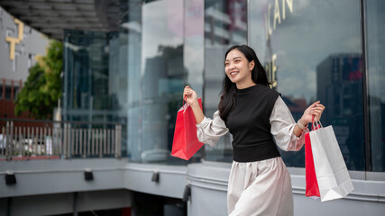 Smiling asian woman or female office worker walking outside shopping mall while carrying paper bags.