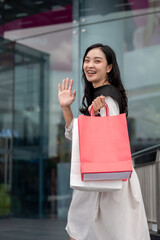 Asian woman office worker waving hand walking into a shopping mall while holding red white paper bag