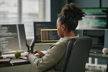 Black young adult woman working at computer desk typing on keyboard, multiple monitors displaying...