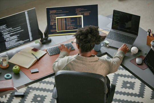 Black young adult woman working at modern desk using desktop computer and laptop, typing code on keyboard, surrounded by multiple monitors, notebooks, and office supplies