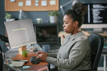 Young adult Black woman working at computer desk typing on keyboard in modern office setting,...