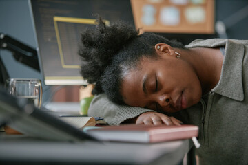Young adult Black woman resting head on desk, appearing asleep in front of computer monitors,...