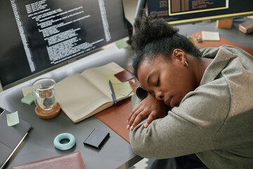 Young adult Black woman sleeping at desk surrounded by computer monitors displaying code, resting...