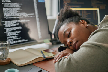 Young adult Black woman sleeping at desk with head resting on arms, surrounded by open notebook,...