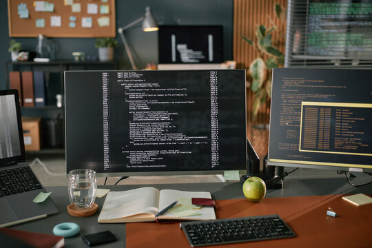 Computer monitor displaying programming code on desk with notebook, pen, glass of water, apple, and sticky notes, suggesting software development or coding workspace in modern office