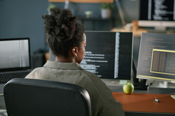 Young Black woman sitting at desk working on computer monitors displaying programming code in...