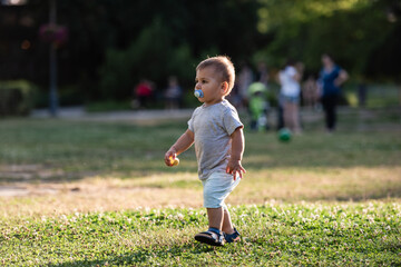 Toddler walking in park with pacifier and fruit snack, enjoying summer day