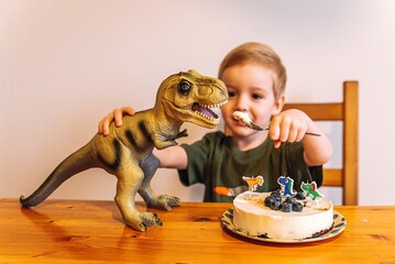 little boy eats cake for birthday 3 years old, plays with toy dinosaur and feeds it with spoons © Julia Suhareva