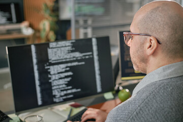 Middle aged Caucasian man working on computer monitor displaying lines of code, sitting at desk in...