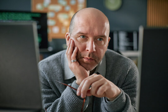 Portrait of middle aged Caucasian man resting chin on hand while sitting at desk, gazing intently at computer monitors, holding eyeglasses, appearing focused on work - Powered by Adobe