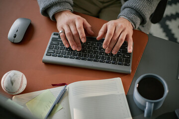 Caucasian middle aged man typing on wireless keyboard at desk with notebook, coffee cup, baseball...
