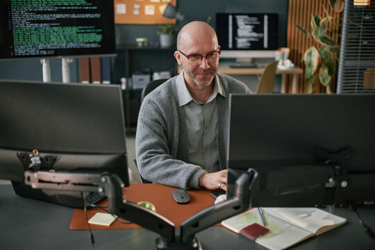 Middle aged Caucasian man sitting at desk working on computer monitors in modern office, typing on keyboard with programming code visible on screens in background