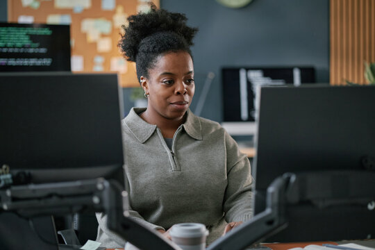 Young adult Black woman working at computer desk in modern office, focusing on dual monitors, coding software visible on screen in background
