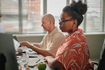 Fototapeta premium Black young adult woman wearing glasses working at desk beside Caucasian man using laptop in modern office setting, both focused on computer screens, natural daylight streaming through windows