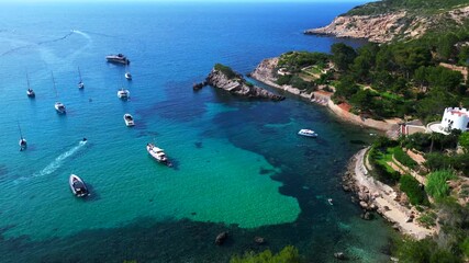 sailboats and yachts moored in turquoise waters of a bay on Ibiza, Spain, surrounded by lush greenery. Marvelous aerial view flight drone top down Above view