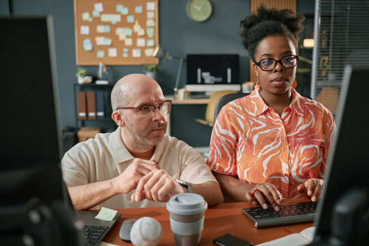 Middle aged Caucasian man wearing glasses sitting beside young adult Black woman typing on computer keyboard, both focusing on computer screens in modern office workspace