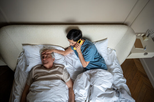 Top view of concerned senior wife checking temperature of sick partner lying in bed while contact employer for time off. Home caregiving, viral infection struggle, compassionate mature woman on phone - Powered by Adobe