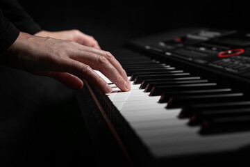 Fototapeta premium A pair of hands with long slender fingers gently presses down on the white keys of a digital piano illuminated by a soft dramatic light against a dark background.