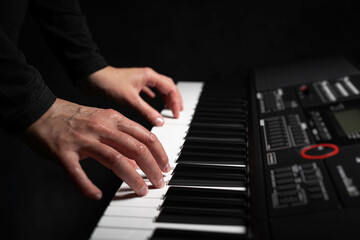 Fototapeta premium A pair of hands with long slender fingers gently presses down on the white keys of a digital piano illuminated by a soft dramatic light against a dark background.