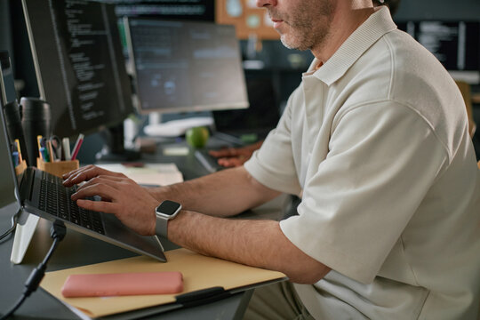 Caucasian middle aged man working on laptop at desk in office, typing on keyboard with smartwatch on wrist, multiple monitors displaying code in background, focused on task
