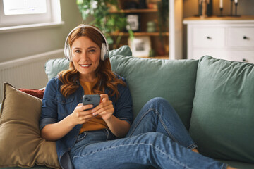 Obraz premium A woman with long red hair sits comfortably on a sofa, wearing headphones and holding a smartphone while smiling and enjoying her leisure time at home.