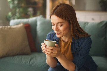 A woman relaxes on a comfortable couch while savoring her warm drink. Soft lighting creates a...