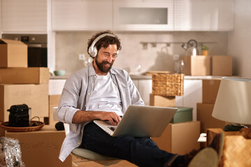 A man is sitting with a laptop on his lap, smiling as he works in a brightly lit living room filled with moving boxes and packing materials, indicating a transition to a new home.