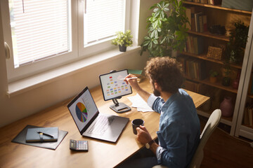 Man focused on work tasks in a cozy home office, utilizing a tablet and laptop while sipping coffee, surrounded by plants and books during daytime hours.