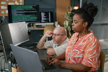 Black young adult woman typing on laptop while Caucasian middle aged man sitting beside her looking...