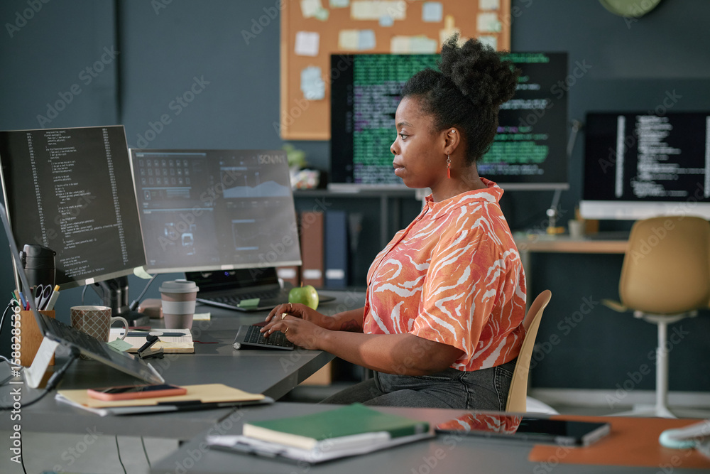 Poster young adult black woman working at computer workstation in modern office, typing on keyboard and foc