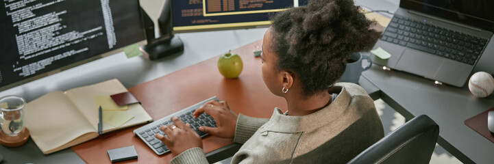 Website header shot of young adult Black woman working at computer desk, typing on keyboard and analyzing code on monitor, surrounded by office supplies and electronic devices in modern workspace