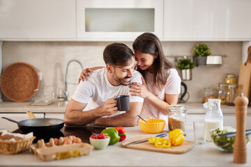 A couple shares a lighthearted moment in their cozy kitchen, surrounded by fresh ingredients, while the man sips a warm drink and the woman leans in with a smile, fostering a joyful atmosphere.