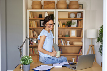 A woman sits on her desk in a cozy home office, smiling while holding a cup of coffee. Bookshelves...