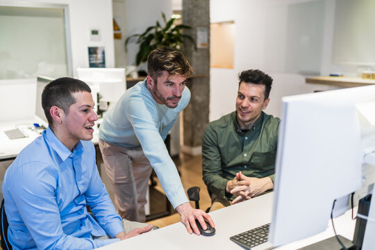 Three young male coworkers cooperating and working together at a desktop computer in a modern coworking office