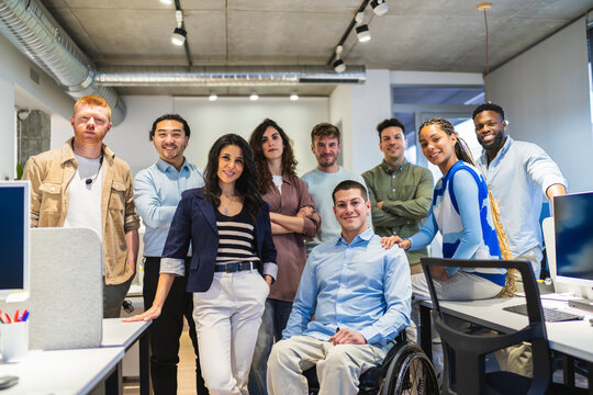Diverse group of professionals standing together in a modern office, showcasing teamwork and inclusion with a colleague using a wheelchair - Powered by Adobe