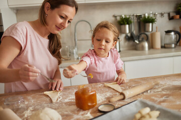 In a bright kitchen, a mother teaches her daughter how to bake. They are covered in flour, smiling while rolling dough and adding preserves to their creations.
