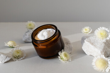 Jar of cosmetic cream on stone table decorated with flowers. Beauty, spa and skincare lifestyle aesthetic concept.