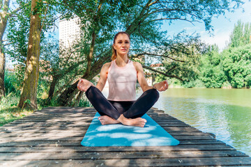 Woman practicing yoga, sitting in lotus pose on wooden pier by urban river, finding tranquility...