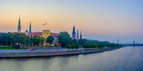 Panoramic view of Riga old town, Latvia at dusk with the Daugava River, Riga Castle, historic church spires, and city skyline under a soft pastel sky.