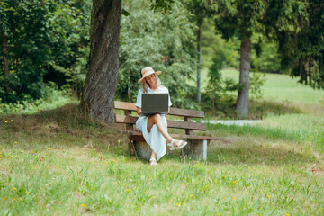 Summer vibes as woman in white dress uses laptop on bench in peaceful rural nature setting
