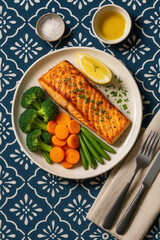 Grilled salmon fillet served with steamed broccoli, carrots, green beans, and lemon wedge on a patterned tablecloth