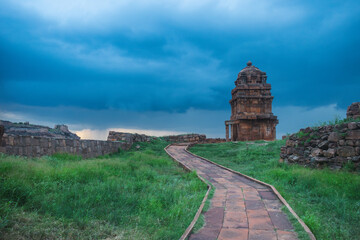 A serene walkway leads to a small ancient temple standing amid lush green grass under a dramatic...