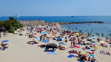 the bustling Plage de la Salis in Antibes on a sunny summer day. The sandy beach is dotted with colorful umbrellas and sunbathers, while people swim and relax in the clear blue Mediterranean Sea.