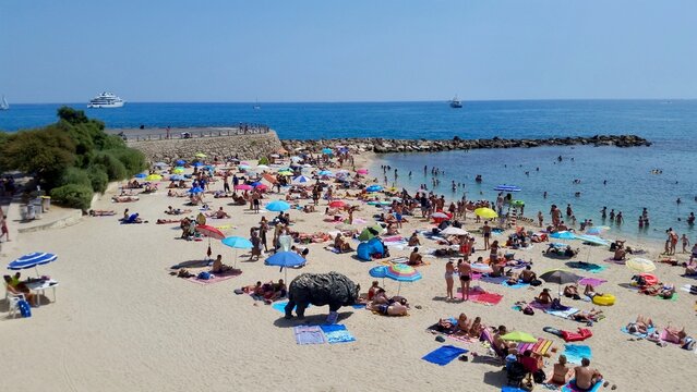 the bustling Plage de la Salis in Antibes on a sunny summer day. The sandy beach is dotted with colorful umbrellas and sunbathers, while people swim and relax in the clear blue Mediterranean Sea.