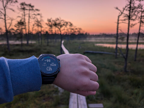 TALLINN, ESTONIA - JULY 17, 2025: A person checks their heart rate and distance on a Garmin Descent Mk3i watch during a morning run on the wooden boardwalk trail at Viru Bog at sunrise.