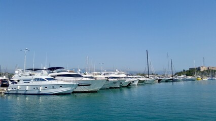 A picturesque view of Antibes' Port Vauban, featuring a line of sleek luxury yachts and sailboats docked in the clear blue water. In the distance, the historic Fort Carré stands on the hillside