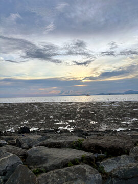A vast muddy tidal flat stretches towards a calm sea under a cloudy sky, with distant land and rocks in the foreground, during a Malaysian evening.