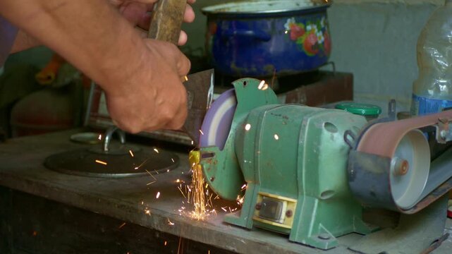 Close-up footage of a man sharpening an axe on a bench grinder in a rustic workshop. Sparks fly as the blade touches the rotating grinding wheel. A raw and authentic view of manual tool maintenance in